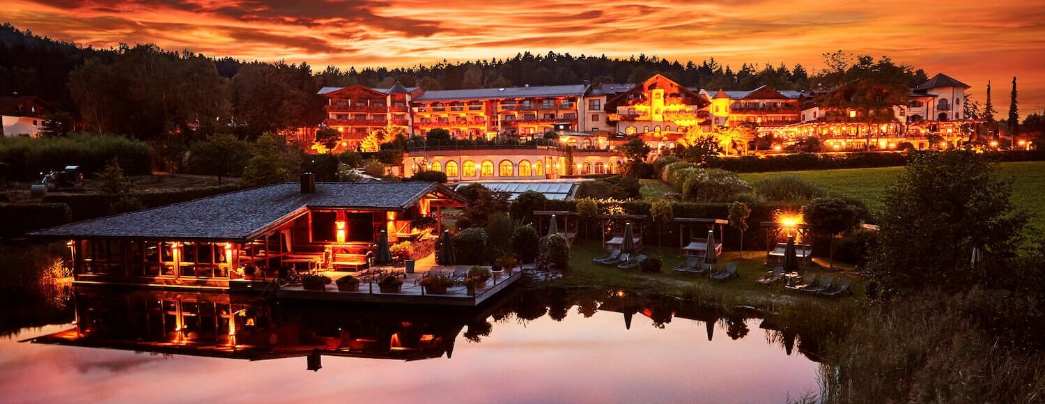 Bayerwaldhof Hotel bei Sonnenuntergang mit warmem Licht, spiegelt sich in einem nahegelegenen Teich, umgeben von Wald und Wiesen.